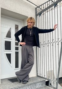 Jeune personne aux cheveux bouclés appuyée contre une grille métallique, portant un pantalon large gris, une chemise noire, une veste noire et des chaussures noires épaisses sur des marches en pierre.