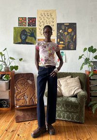 Camisa de manga curta com padrão floral, calças de perna larga em denim escuro, sapatos de plataforma castanhos, com um sofá verde texturizado e arte de parede ao fundo.