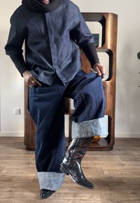 Personne portant un jean en denim foncé avec larges revers, un t-shirt à manches longues noir sous une chemise en denim à manches courtes, et des bottes de cowboy noires, levant une jambe en intérieur.