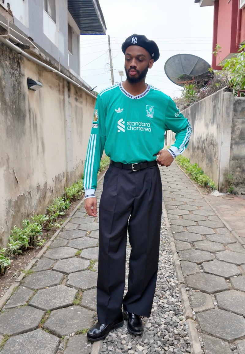 Maillot de gardien de but du Liverpool FC vert avec des bandes blanches, pantalon noir et béret noir. Chemin pavé en pierre et végétation en arrière-plan.