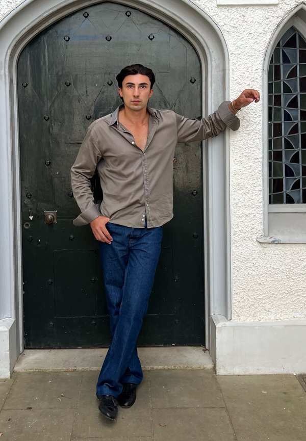 Homme aux cheveux foncés portant une chemise grise et un jean bleu appuyé contre une porte voûtée sombre dans un mur blanc texturé avec une fenêtre voûtée.