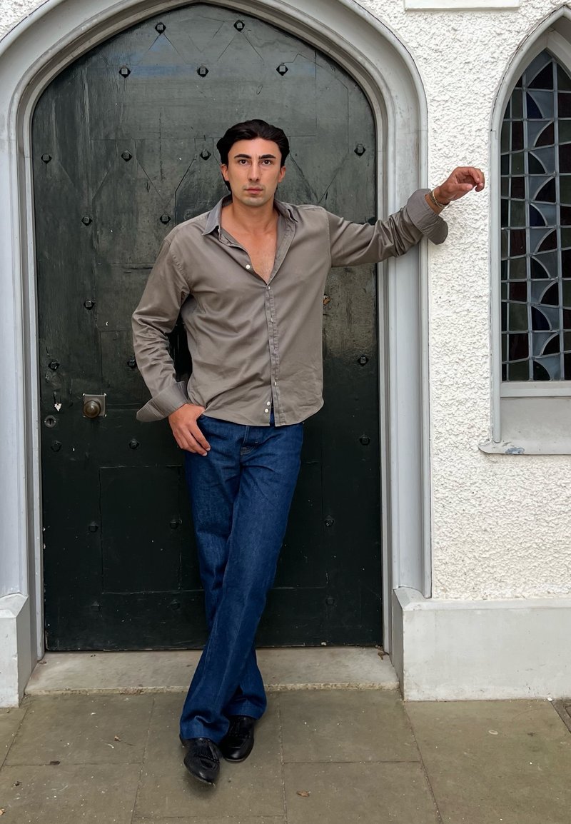 Homme aux cheveux foncés portant une chemise grise et un jean bleu appuyé contre une porte voûtée sombre dans un mur blanc texturé avec une fenêtre voûtée.