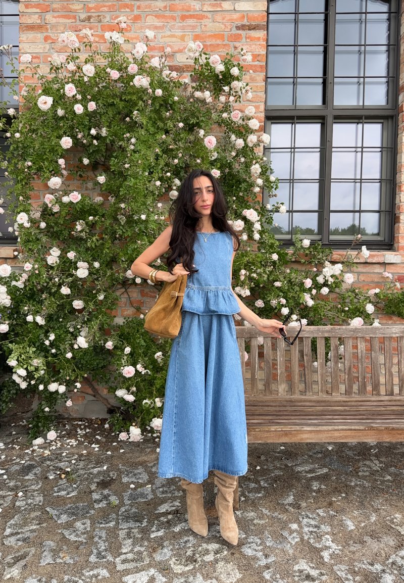 Femme en haut en denim et jean à jambes larges debout près d'un banc en bois et d'un buisson de roses roses en fleurs contre un mur en briques avec de grandes fenêtres.