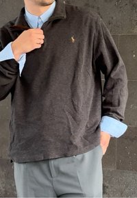 Mann, der den Kragen eines dunkelgrauen Pullovers über einem hellblauen Hemd anpasst, trägt graue Hosen und steht vor einem Hintergrund aus dunklen Fliesen.