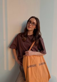 Ruskea ylisuuri t-paita, oranssi kangaskassi, jossa on "SAM SøE" -logo paksulla kirjaimella, pyöreät silmälasit, neutraali tausta lämpimällä valaistuksella.