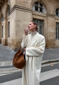 Une femme en manteau de laine blanc et col roulé beige applique du rouge à lèvres. Elle tient un sac à main en daim marron. L'arrière-plan présente une architecture en pierre.