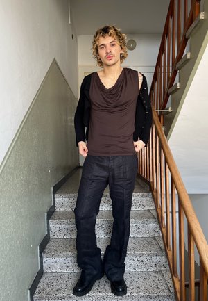 Persona con cabello rizado y rubio de pie en unas escaleras interiores, vestida con una blusa marrón sin mangas y drapeada, pantalones negros y zapatos negros, sosteniendo una chaqueta negra.