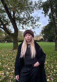 Osoba v tmavom kabáte a hnedej kožušinovej čiapke stojí v parku s trávou a padlými listami. Viditeľné sú dlhé rovné vlasy a náhrdelník.