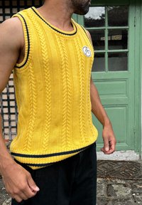 Jersey sin mangas de color amarillo con patrón de trenzas, acento azul marino en el cuello y el dobladillo, parche redondo con logo, usado sobre pantalones oscuros.