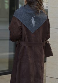 Manteau en daim marron avec un large col en laine grise arborant un logo. Le manteau a une ceinture et arrive jusqu'au milieu du mollet, avec une texture lisse.