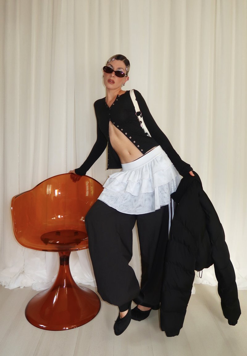 Personne portant un cardigan noir court, une jupe blanche superposée sur un pantalon large noir, tenant une veste noire, appuyée sur une chaise translucide orange.