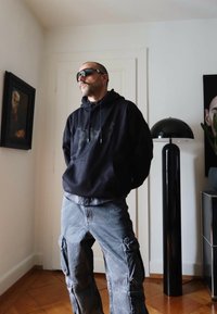 Felpa nera con cappuccio, pantaloni cargo grigi con multiple tasche, occhiali da sole. Stanza con lampada moderna e arte astratta sulla parete.