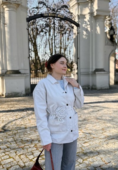 Joven mujer con chaqueta blanca con detalles de flores, de pie junto a una puerta negra ornamentada y columnas de piedra, sosteniendo un bolso rojo a la luz del día.