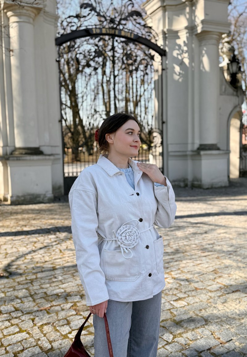 Nuori nainen valkoisessa takissa, jossa on kukkakoriste, seisoo koristeellisen mustan portin ja kivipylväiden vieressä pitäen punaista laukkua päivänvalossa.