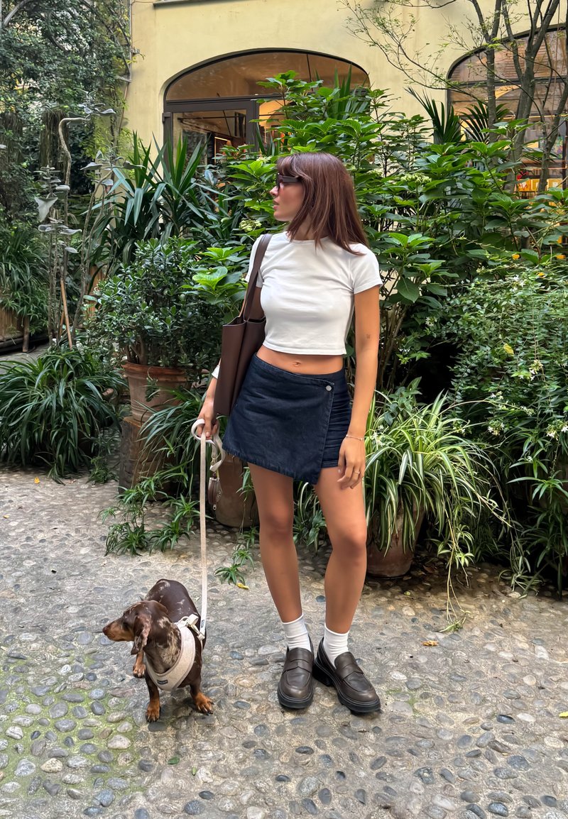 Donna in un crop top bianco e una gonna blu che tiene un guinzaglio, in piedi accanto a un bassotto marrone su un sentiero di pietra, con piante verdi sullo sfondo.