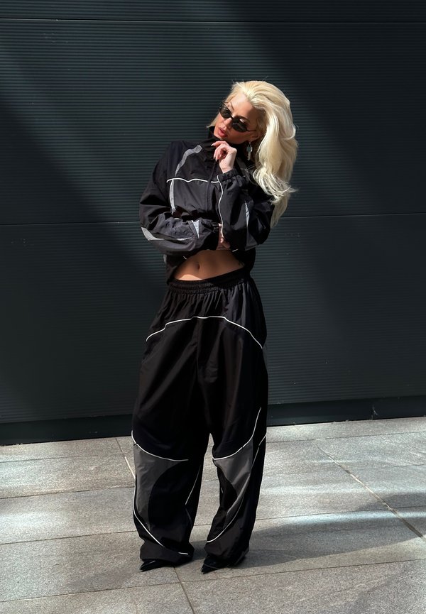 Femme blonde en pantalon et veste noirs oversize avec des détails réfléchissants pose la main près du menton devant un mur sombre.