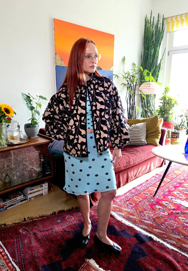 Mujer con cabello largo y rojo y gafas, lleva una chaqueta estampada sobre un vestido azul con lunares, de pie en una sala de estar colorida con plantas y alfombras.