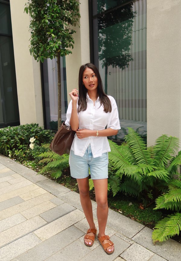 Donna con camicia bianca e pantaloncini di jeans si trova su un marciapiede urbano vicino a piante verdi, con una grande borsa marrone sulla spalla.
