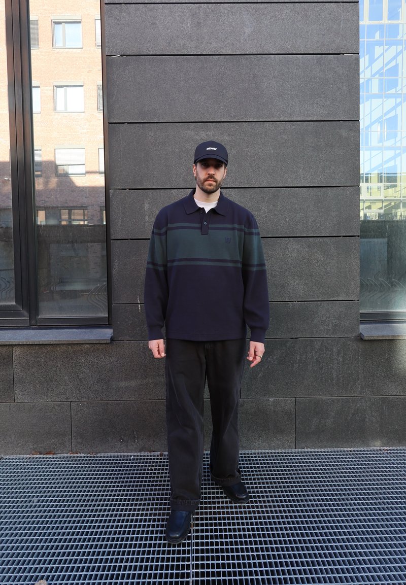Homme portant une casquette noire, un pull bleu marine à rayures vertes, un pantalon noir et des chaussures noires, debout contre un mur en pierre sombre avec des fenêtres.