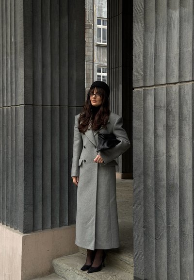 Abrigo gris cruzado con bolsillo de solapa, combinado con un bolso de mano negro. Cabello largo, gafas y sombrero negro; de pie junto a columnas grises con textura.