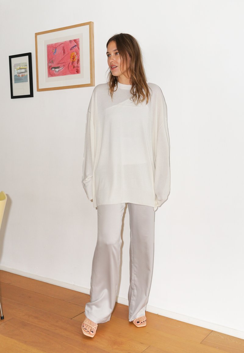 Yliösikoinen kerma sävyinen neule, jossa on teksturoitu pinta, yhdistettynä vaaleanharmaisiin satiinilahkeisiin. Mallilla on päällään ruskeat korkokengät. Taustalla valkoinen seinä.