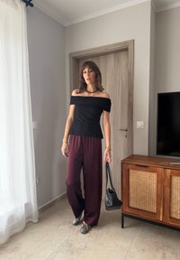 Sieviete melnā topā ar atklātām pleciem un vaļīgās sarkanbrūnās biksēs, tur melnu rokas somiņu, stāv modernā telpā pie koka skapja.