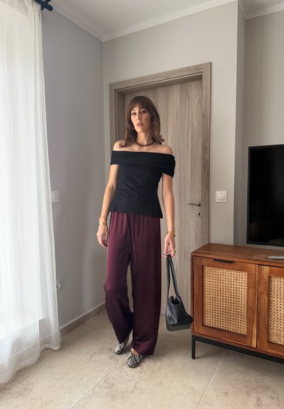 Mujer con top negro de hombros descubiertos y pantalones sueltos color granate, sosteniendo un bolso negro, de pie en una habitación moderna cerca de un mueble de madera.