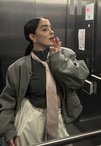 Šedá přepálená bomber bunda, těsný tmavý svetr, bílé plisované kalhoty, růžová texturovaná kravata, kulaté náušnice, v výtahu.