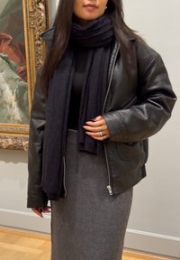 Crna kožna jakna s oversized krojem, uparena s sivom rebrastom midi suknjom i odgovarajućim crnim šalom. Ruke su vidljive, prikazujući prstenje.