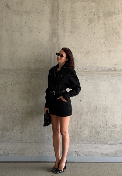 Mujer que lleva un vestido vaquero negro con cinturón, tacones negros y gafas de sol, de pie junto a una pared de hormigón gris texturizada sosteniendo un bolso de mano.