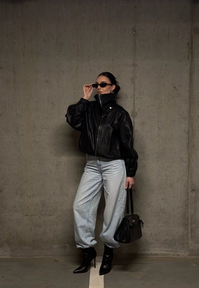Mujer con chaqueta de cuero negra, jeans claros, botas negras de tacón, gafas de sol y bolso negro, de pie contra una pared de cemento.