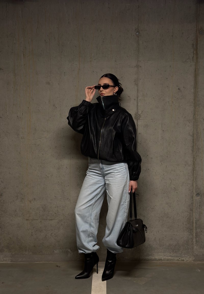 Donna con giacca di pelle nera, jeans chiari, stivali neri con tacco, occhiali da sole e borsa nera, in piedi contro un muro di cemento.
