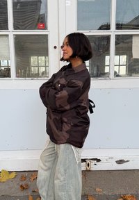 Hnedá maskovacia bunda s voľným strihom, veľkým predným vreckom a nastaviteľným šnúrkou na zadnej strane, v kombinácii s ľahkými, vrstvenými nohavicami.