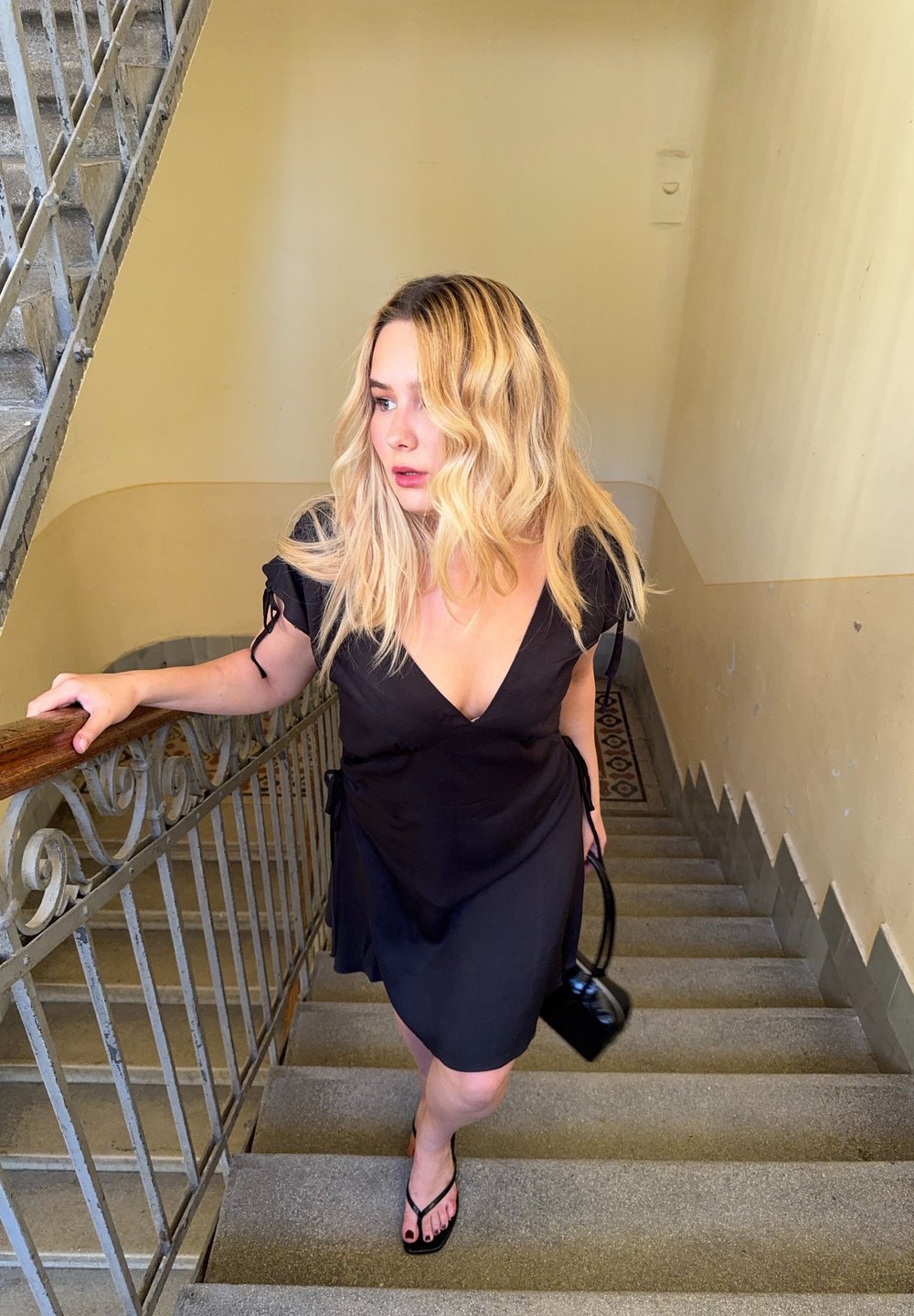 Vestido preto envolvente com mangas curtas, laços discretos na cintura e um decote em V profundo. Usado com sandálias pretas planas, segurando uma pequena bolsa preta.