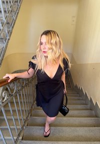 Vestido preto envolvente com mangas curtas, laços discretos na cintura e um decote em V profundo. Usado com sandálias pretas planas, segurando uma pequena bolsa preta.