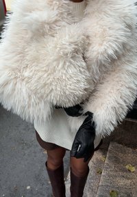 Pluizige, gebroken-witte nep-bont wikkel met een dikke textuur en een hoge kraag, gecombineerd met zwarte leren handschoenen en een witte trui eronder.