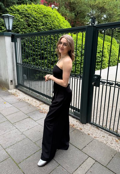 Top negro sin mangas combinado con pantalones negros holgados y zapatos blancos; la escena se ambienta cerca de un seto verde y una verja.