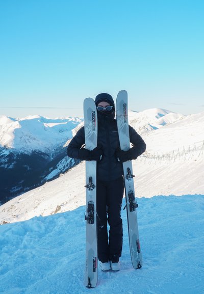 Persona con equipo de invierno negro y gafas de sol sosteniendo dos esquís verticales en una montaña nevada con un cielo azul claro de fondo.