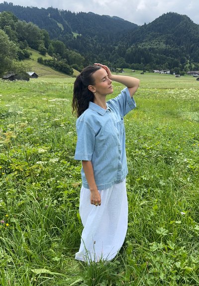 Camisa de mezclilla, cuello, mangas cortas, azul claro; combinada con una falda larga blanca. De pie en un campo de hierba con árboles y montañas al fondo.