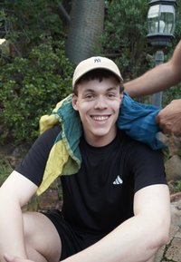 Jeune homme portant une casquette et une tenue de sport noire sourit pendant qu'une autre personne lui drape une serviette jaune et bleue sur les épaules en plein air.