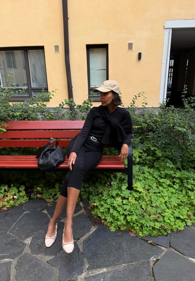 Blusa negra de manga larga, pantalones negros de tiro alto, gorra beige y zapatos rosa claro. Bolso negro sobre un banco rojo rodeado de follaje verde.