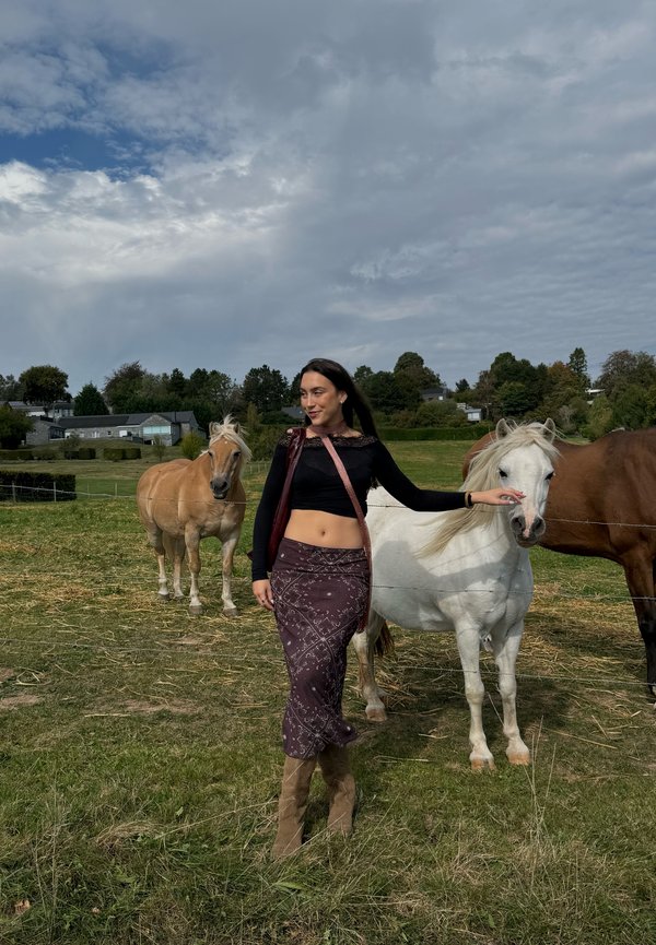 Mujer de pie en un campo de hierba acariciando a un caballo blanco, con otros caballos y casas visibles bajo un cielo parcialmente nublado.