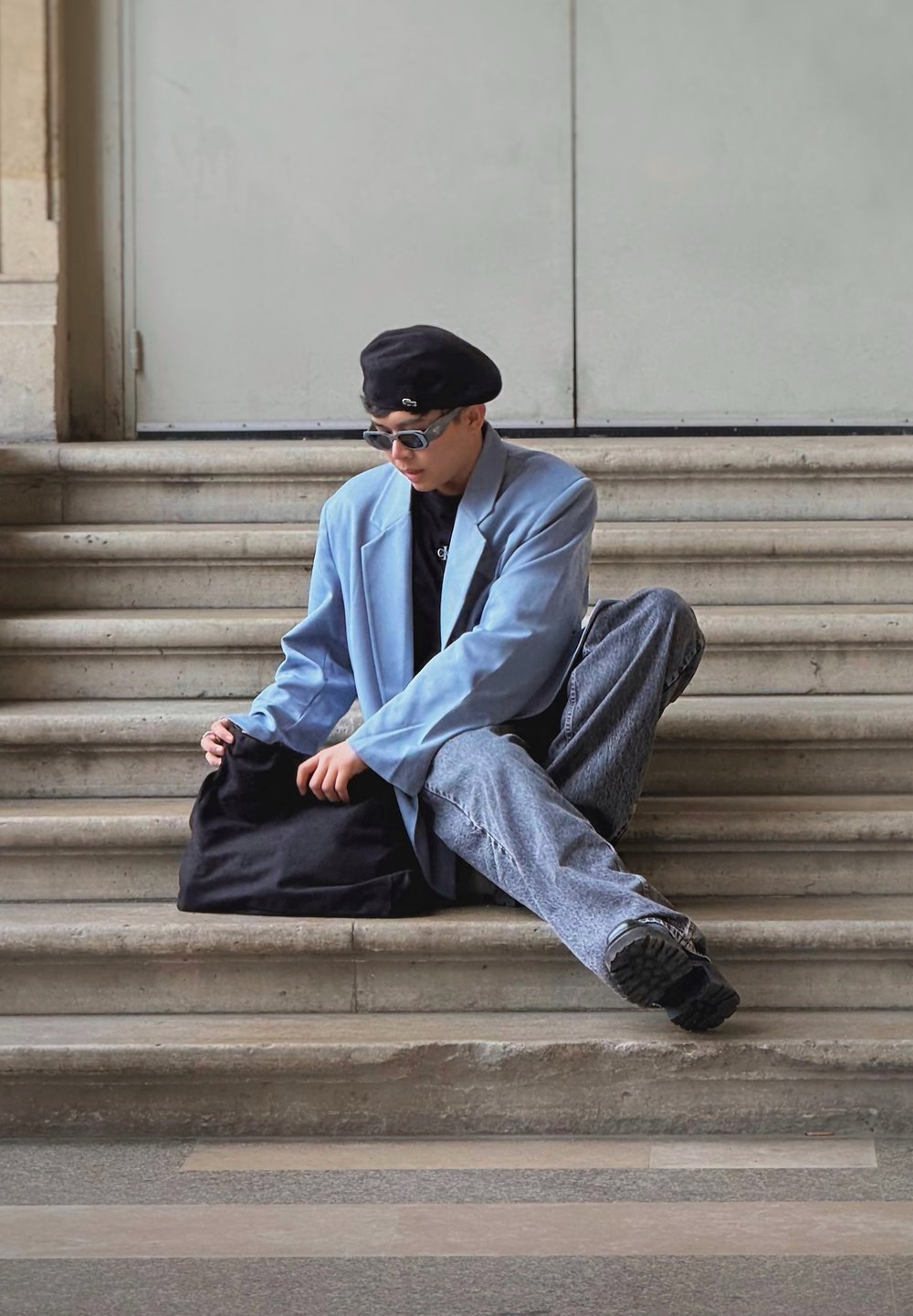 Heledad sinine ülikond, must kott, must särk, prillid, lõdvad hallid teksad, istudes kiviastmetel neutraalse tausta ees.