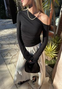 Blusa preta de manga longa com textura franzida, combinada com uma saia creme. A segurar uma mala de mão de vime preta e a usar sapatos com padrão de vaca.