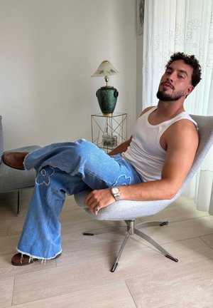 Pantalones de mezclilla acampanados de un azul claro con bordado floral, combinados con una camiseta blanca. Sentada en una silla gris, con zapatos marrones visibles. Decoración minimalista.