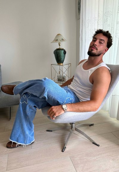 Pantalones de mezclilla acampanados de un azul claro con bordado floral, combinados con una camiseta blanca. Sentada en una silla gris, con zapatos marrones visibles. Decoración minimalista.