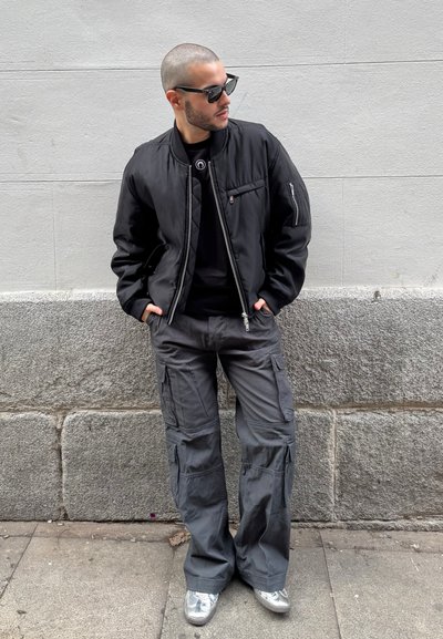 Chaqueta bomber negra con bolsillos con cremallera, camiseta negra, pantalones cargo grises con bolsillos laterales y zapatillas plateadas.
