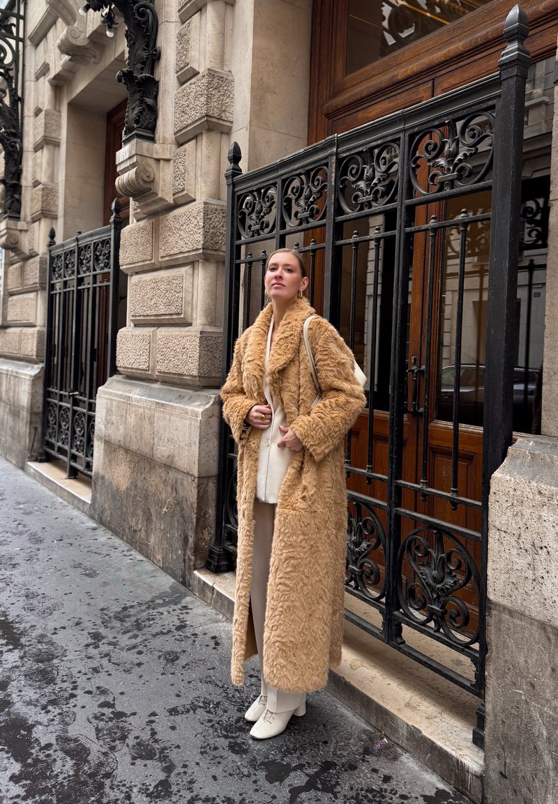 Mujer con abrigo largo de piel color marrón claro, pantalones blancos y zapatos blancos, de pie sobre una acera mojada de la ciudad junto a una ornamentada puerta negra de hierro forjado y un edificio de piedra.