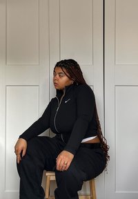 Μαύρη φανέλα με φερμουάρ και μακριά μανίκια, με ριγωτή υφή, συνδυασμένη με χαλαρούς μαύρους παντελόνες. Μοντέλο καθισμένο σε σκαμπό μπροστά από φωτεινό φόντο.