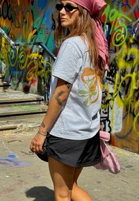 Biela tričko s farebným grafickým dizajnom, spárované s čiernym šortkami. Doplnky zahŕňajú ružovú tašku a slnečné okuliare. Mestský graffiti pozadie.